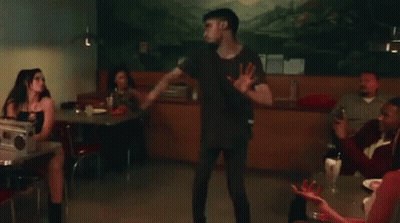 A Group Of People Are Sitting At Tables In A Diner With A Man Standing In The Background . GIF