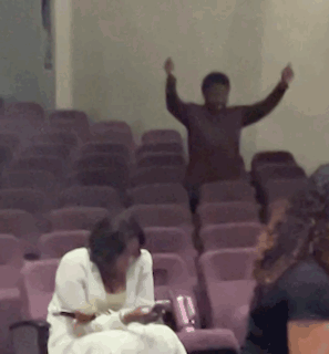 A Group Of People Are Sitting In A Row Of Purple Chairs With Their Arms In The Air GIF