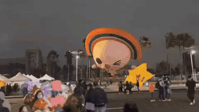 A Group Of People Are Standing Around A Balloon That Looks Like A Cat Flying In The Air . GIF