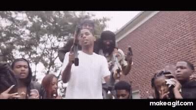 A Group Of People Are Standing Around A Man Holding A Gun In Front Of A Brick Building . GIF
