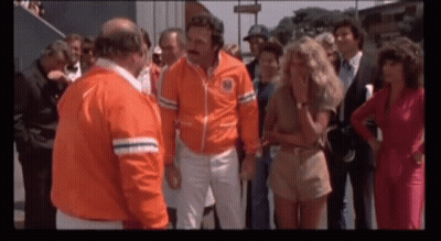 A Group Of People Are Standing Around A Man Wearing An Orange Jacket That Says ' New York ' On It GIF
