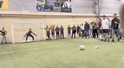 A Group Of People Are Standing Around A Soccer Field Watching A Goalie Kick A Soccer Ball . GIF