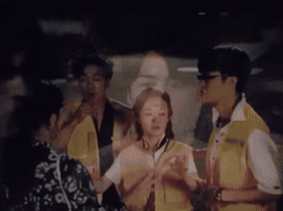 A Group Of People Are Standing In A Dark Room With A Man 'S Face Projected On A Wall Behind Them . GIF