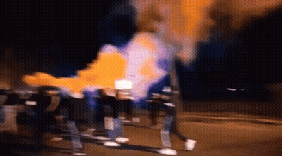 A Group Of People Are Walking Down A Street At Night With Smoke Coming Out Of A Fire . GIF