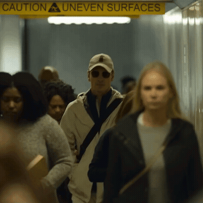 A Group Of People Are Walking In A Hallway Under A Yellow Sign That Says Caution Uneven Surfaces GIF