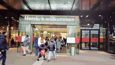 A Group Of People Are Walking In Front Of A Store That Says Herzlich Willkommen GIF