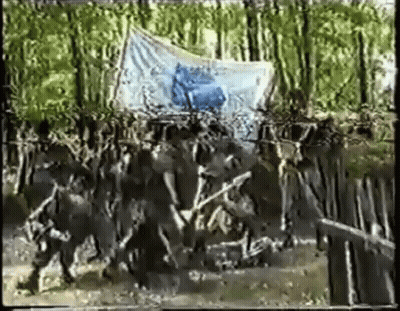 A Group Of People Are Walking In The Woods With A Large Flag In The Background . GIF