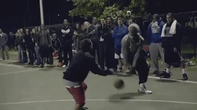 A Group Of People Are Watching Two Men Play Basketball On A Court At Night GIF