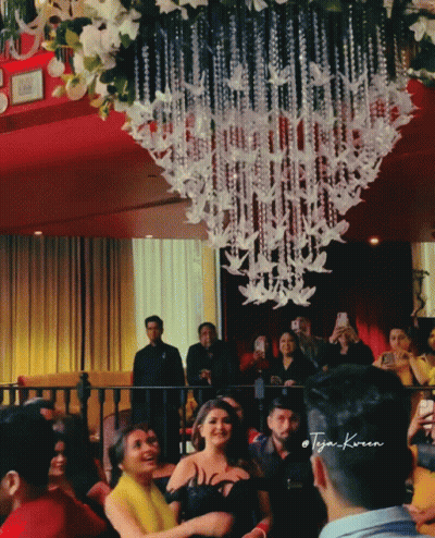 A Group Of People Gathered In A Room With A Chandelier Hanging From The Ceiling And A Man Taking A Picture GIF
