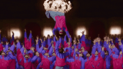 A Group Of People In Pink And Blue Jackets Are Standing In A Crowd And A Person Is Being Thrown In The Air . GIF