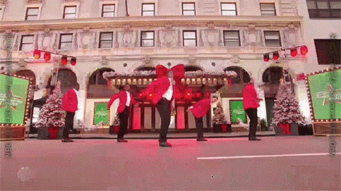 A Group Of People In Red Jackets Are Dancing In Front Of A Building With Christmas Trees . GIF