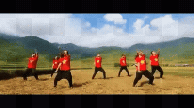 A Group Of People In Red Shirts Are Dancing In A Field With Mountains In The Background . GIF