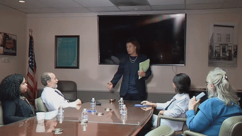A Group Of People Sitting Around A Conference Table With A Sign On The Wall That Says ' Sidney Smith ' GIF