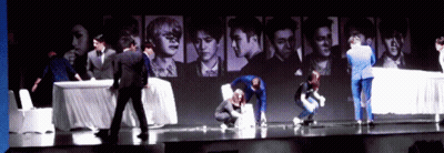 A Group Of People Standing On A Stage In Front Of A Wall With Pictures Of People GIF