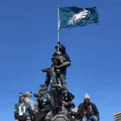 A Group Of People Standing On Top Of A Statue Holding A Flag With The Eagles Logo On It GIF