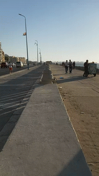 A Group Of People Walking Down A Sidewalk With A Trash Can On The Side GIF