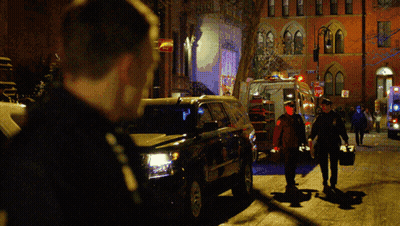 A Group Of People Walking Down A Street At Night With An Ambulance Behind Them GIF