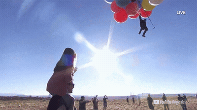 A Group Of People Watching A Man Flying Through The Air With Balloons Attached To Him GIF