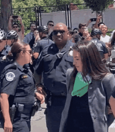 A Group Of Police Officers Are Standing Around A Woman Wearing A Green Scarf GIF