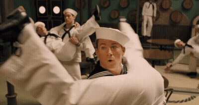 A Group Of Sailors Are Holding A Man In Their Arms In A Room With Barrels On The Wall GIF