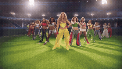 A Group Of Women Are Dancing On A Field With One Wearing A Yellow Outfit GIF