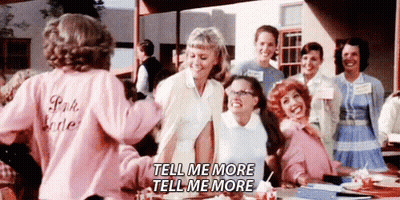 A Group Of Women Are Standing Around A Table Talking To Each Other . One Of The Women Is Wearing A Pink Shirt . GIF