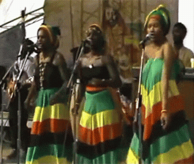 A Group Of Women Singing Into Microphones While Wearing Colorful Skirts GIF
