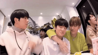 A Group Of Young Men Are Sitting At A Table And One Of Them Is Hugging Another Man . GIF