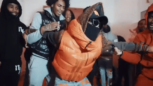 A Group Of Young Men Are Standing Around A Man In An Orange Jacket . GIF