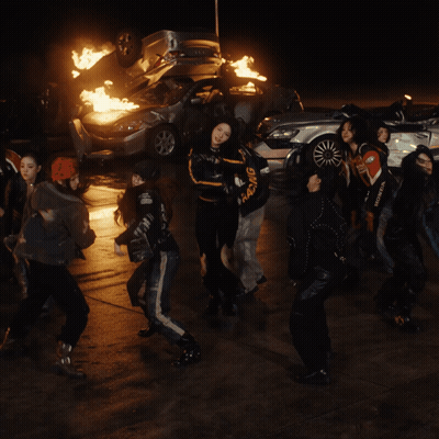 A Group Of Young Women Are Dancing In Front Of A Pile Of Burning Cars GIF