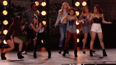 A Group Of Young Women Are Dancing On A Stage With A Man Holding A Microphone GIF
