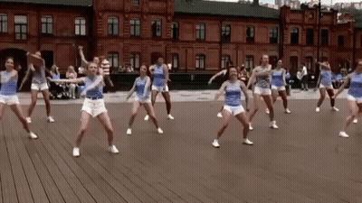 A Group Of Young Women Are Dancing On A Wooden Floor In Front Of A Brick Building . GIF