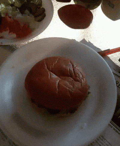A Hamburger On A White Plate With A Newspaper On The Table In The Background GIF