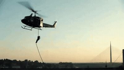 A Helicopter With A Rope Attached To It Is Flying Over A Bridge GIF