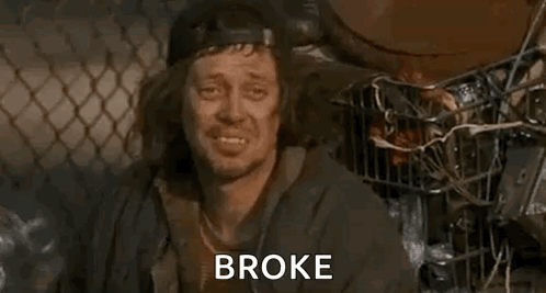 A Homeless Man Is Sitting In Front Of A Chain Link Fence With The Words `` Broke '' Written On His Face . GIF