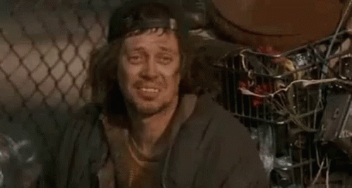 A Homeless Man Wearing A Baseball Cap And A Jacket Is Smiling While Sitting In Front Of A Chain Link Fence . GIF
