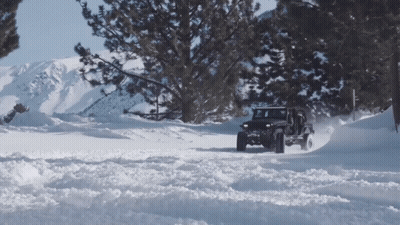 A Jeep Is Driving Through A Snowy Forest With Mountains In The Background GIF