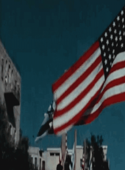 A Large American Flag Is Flying In The Wind In Front Of A Building GIF