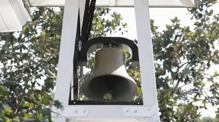 A Large Bell Is Hanging From A White Structure With Trees In The Background GIF