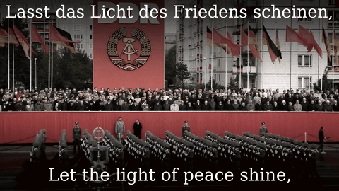 A Large Crowd Of People Are Gathered In Front Of A Large Red Banner That Says Ddr Let The Light Of Peace Shine GIF
