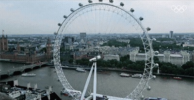 A Large Ferris Wheel In The Middle Of A City With The Olympics Logo On The Bottom GIF