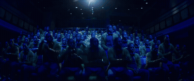 A Large Group Of People Are Sitting In A Dark Auditorium Watching A Movie GIF
