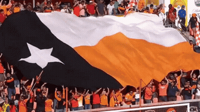 A Large Orange And White Flag With A White Star On It Is Being Held Up By A Crowd GIF