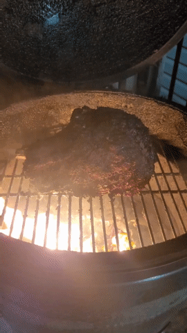 A Large Piece Of Meat Is Cooking On A Grill With Flames Behind It GIF