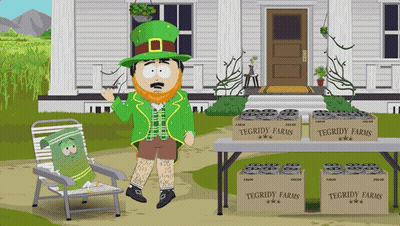 A Leprechaun Stands In Front Of A Table With Boxes Of Tegridy Farms On It GIF