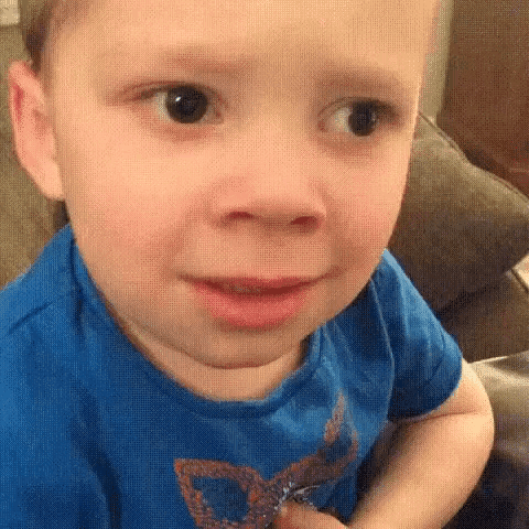 A Little Boy In A Blue Shirt Is Sitting On A Couch Making A Funny Face . GIF