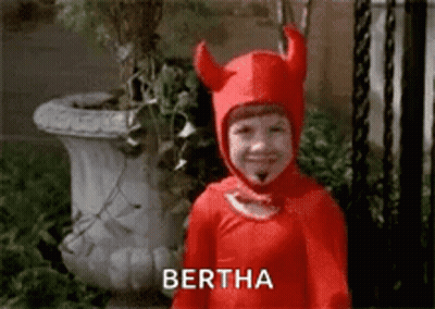A Little Boy In A Red Devil Costume Is Smiling And Standing In Front Of A Potted Plant . GIF