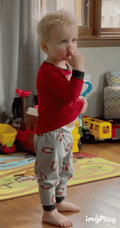 A Little Boy In A Red Shirt And Gray Pants Is Standing In A Living Room With Toys . GIF
