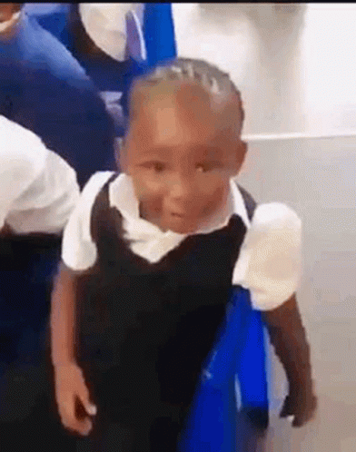 A Little Boy In A School Uniform Is Standing Next To A Blue Chair In A Classroom . GIF