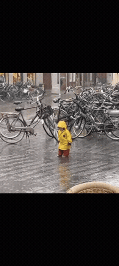 A Little Boy In A Yellow Raincoat Is Standing In The Rain In Front Of A Row Of Bikes . GIF